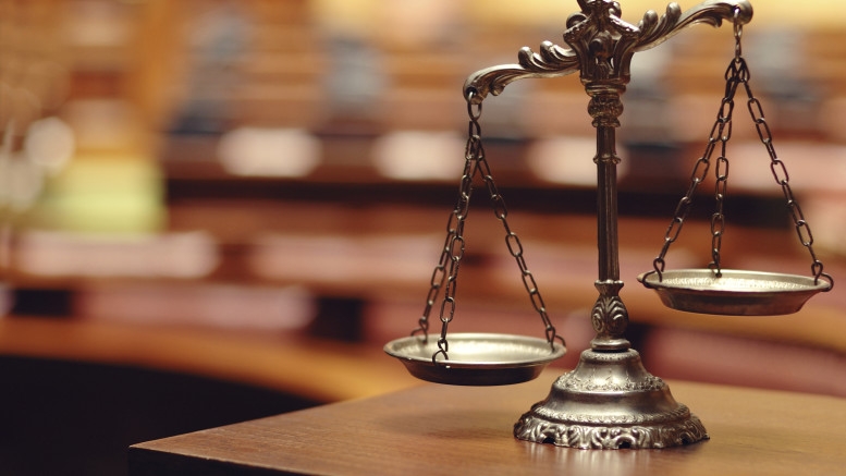 A photo of an old brass set of scales, sitting on a wooden desk, with a blurry courtroom in the background.