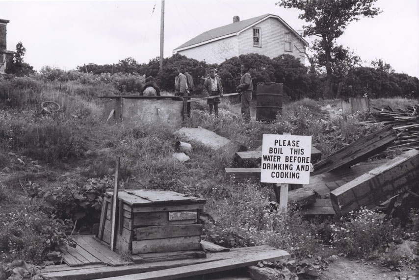 A black and white photo of a community with a house in the background and three men standing along a wall. In the foreground of the photo is a sign next to a well that says please boil the water before drinking and cooking. 