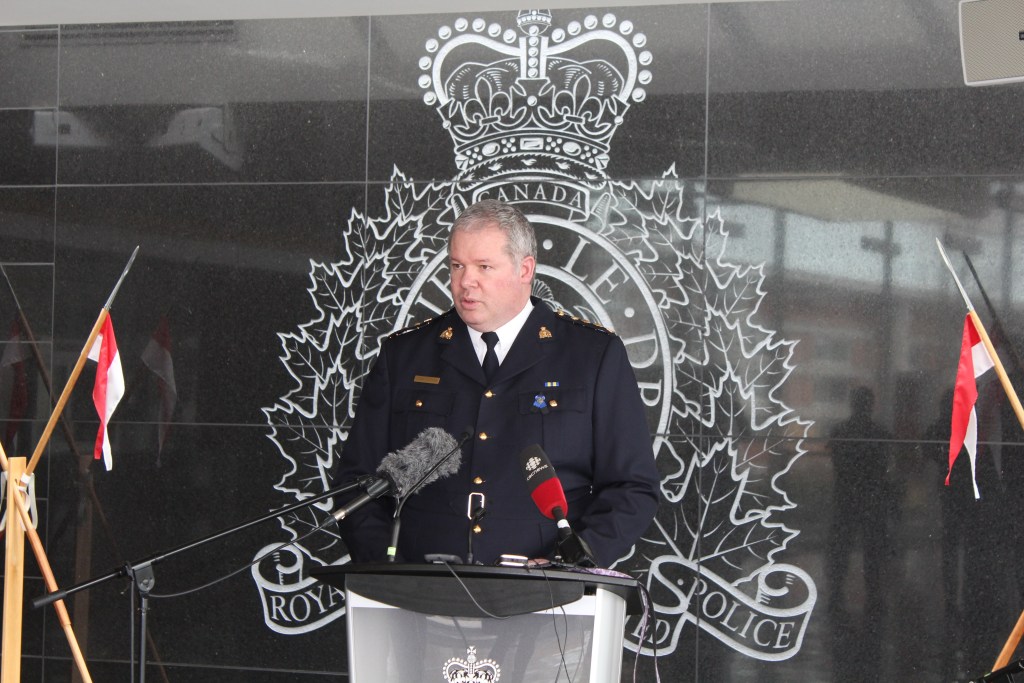 A white middle aged man in a black RCMP uniform speaks in front of a polished granite wall with the RCMP crest etched into it.