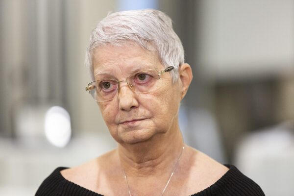 A photo of white woman with short silver hair, glasses, black wide-neck sweater, and long silver chain.
