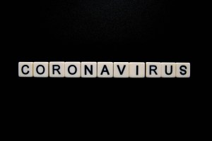 This is a photo of white letter tiles on a black backgound. The black text on the letters spells out coronavirus in capital letters.