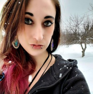 A selfie of Ashleen Louise D'Orsay, taken outside on a snowy day. she's wearing a black hoodie and dramatic makeup. she has some face piercings and cool turquoise pendant earrings, and her long brown hair has some great red colour on the ends.