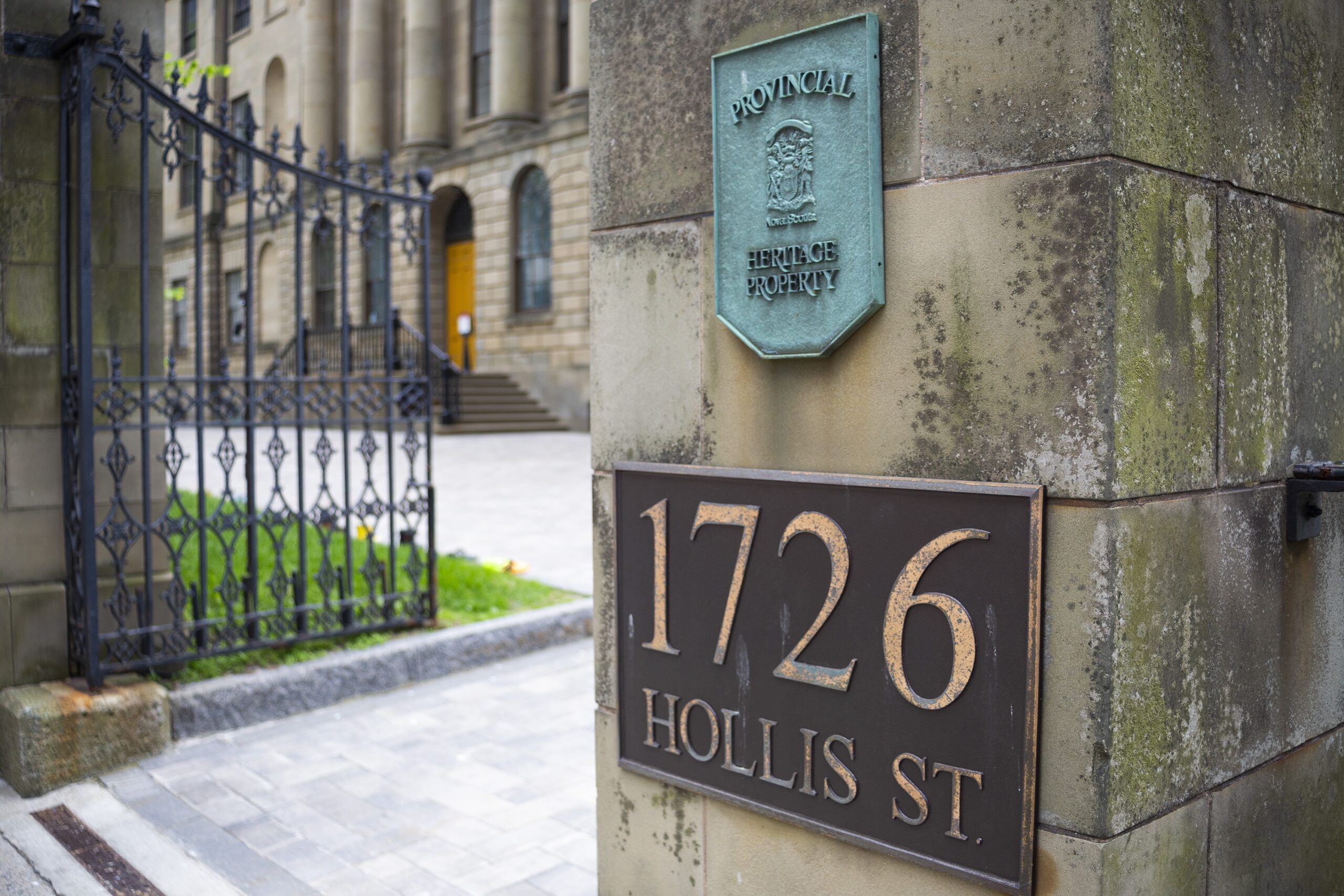 The open wrought iron entrance gate to the courtyard of Province House in June 2021. On the stone wall is a bronze plaque reading 1726 Hollis St, and above that a copper plaque, completely green with patina, designating the building a provincial heritage property.