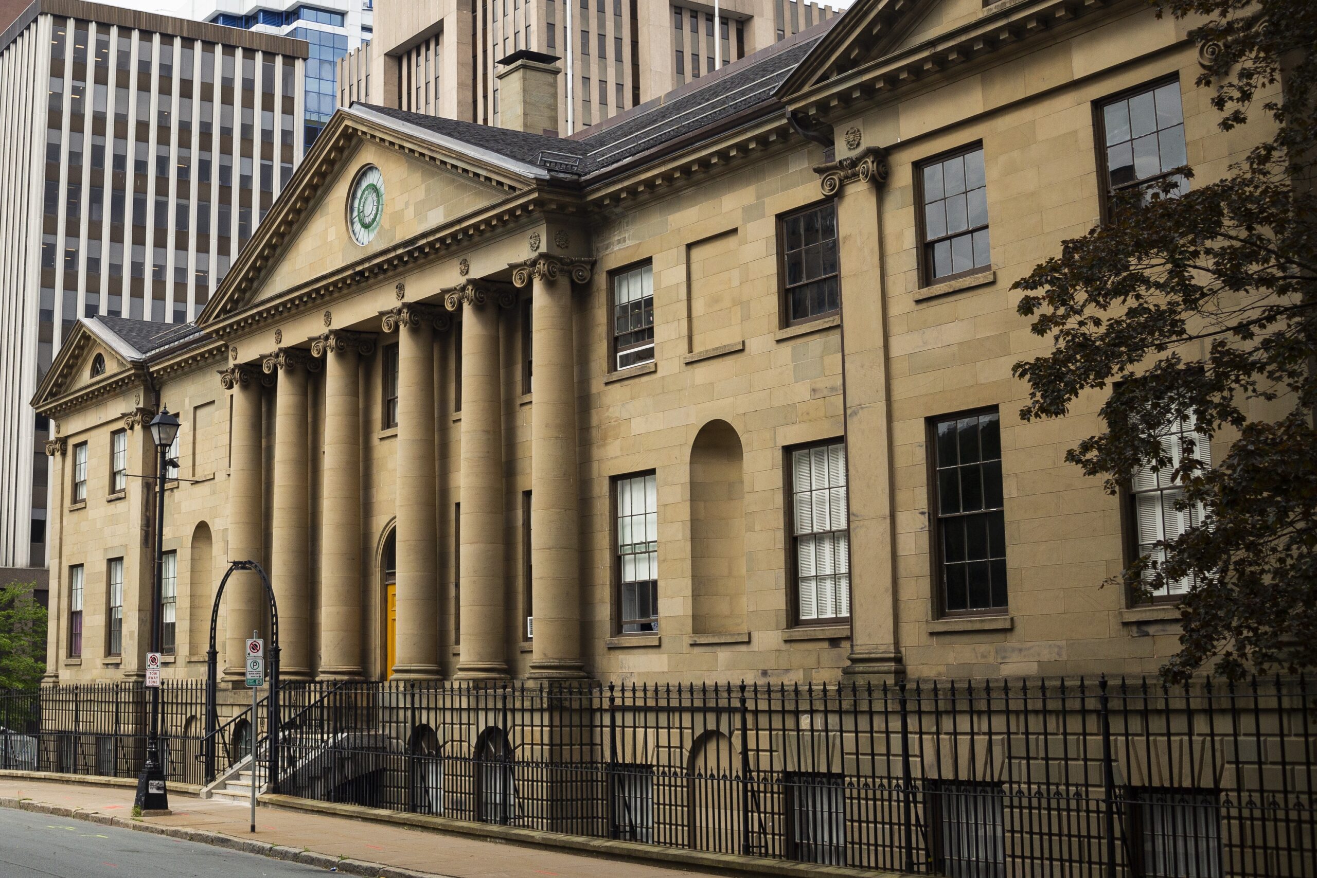 The front of Province House in June 2021. In the front is a very clean sidewalk and wrought iron fence; in the background, rising high above the roofline, are more modern buildings.