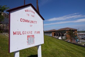 A white sign shaped like a house with a peaked roof on which is written Welcome to the community of Mulgrave Park. Below that is a logo of three people in grey on a red square. The red paint has faded in the sun. In the background you can see the slope of the grassy lawn, a chain link fence, several homes in a row of townhouses, and beyond that Barrington Street. Taken in 2018.