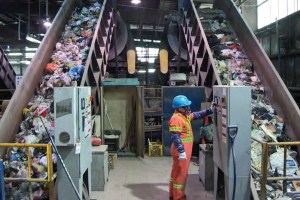 Two conveyer belts filled with garbage are seen sloping up toward the ceiling in an industrial setting. In the centre-right of the frame, a worker wearing a high-visibility orange suit, blue hard hat, and respirator mask operates the conveyer belt to the right.