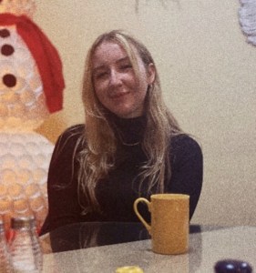 Lauren Petrie. she has long blonde hair and a black turtleneck sweater. She's smiling at the camera, sitting at a table in a restaurant or cafe.