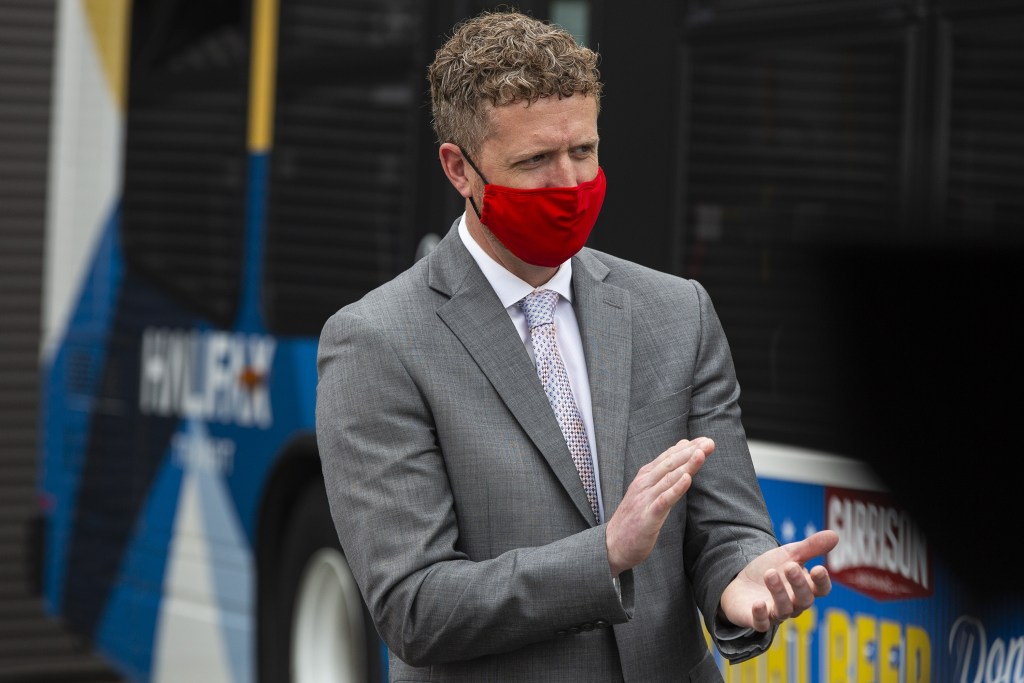 A man wearing a grey suit, white shirt, dotted tie and a red mask claps while standing in front of a bus.