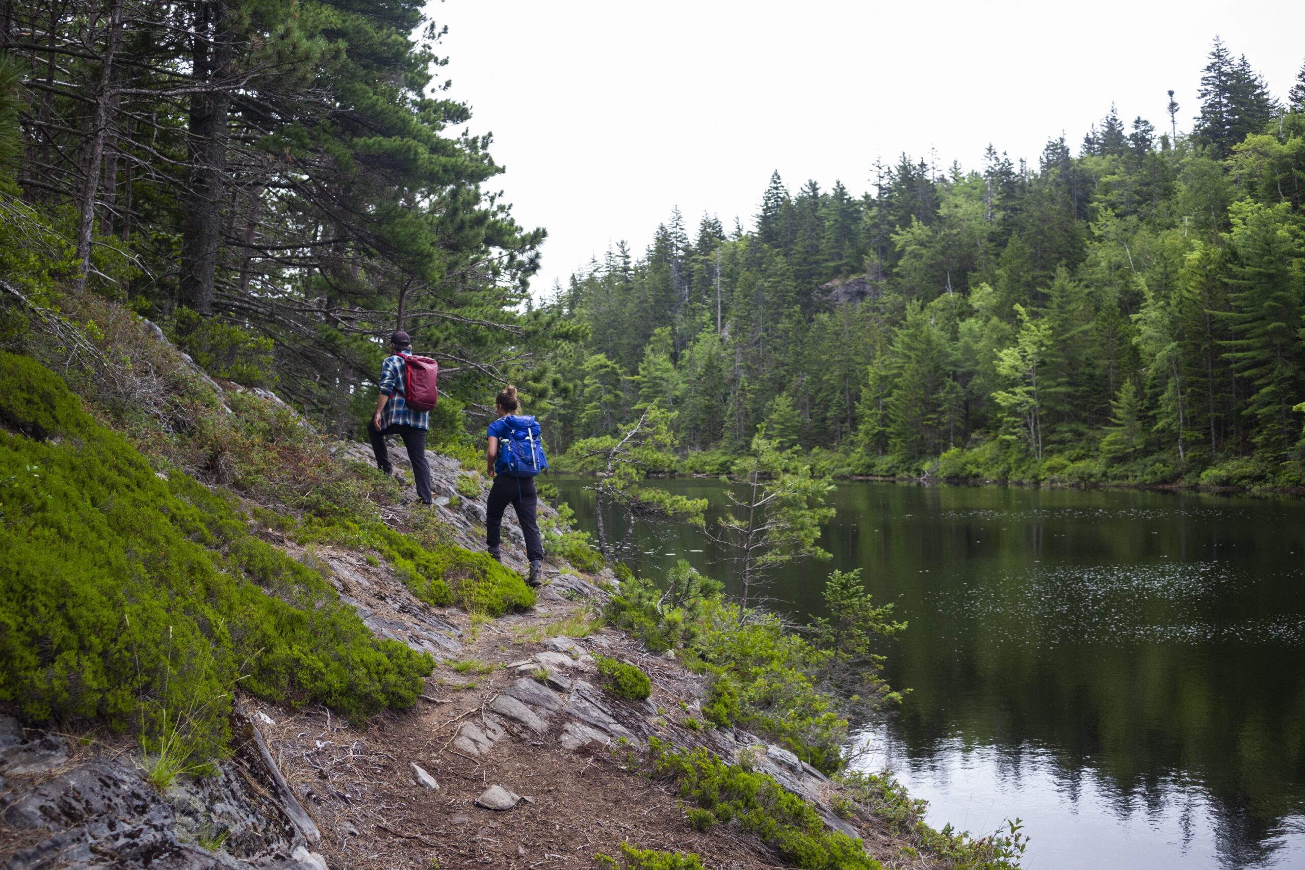 Two people hike along a lake. On the right is a woman with a blue backpack, and on the left, in front, is a man with a red backpack. It's an overcast day, and along the shores of the lake there are evergreen trees and steep hills.