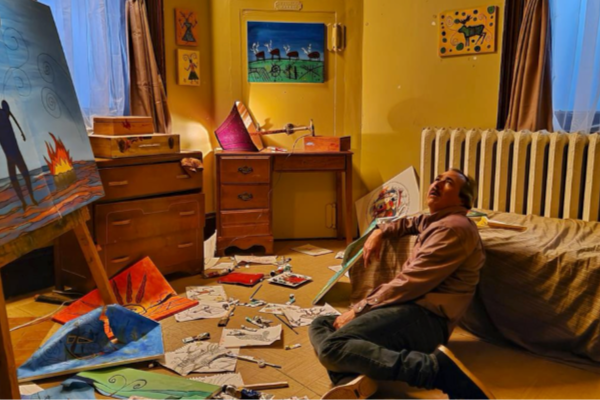 A man sits in anguish on the floor of a bedroom with an artist's easel, and there are handpainted pictures scattered on the floor