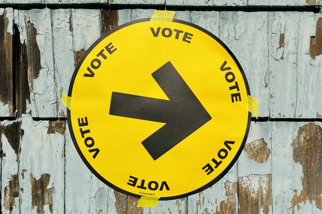 A circular yellow sign with a black arrow and the word VOTE printed six times around the circle is seen taped to weathered blue-green cedar shingles.