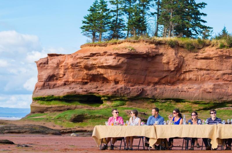 People sitting at a table with a linen table cloth set up on a beach at low tide, with a dramatic stone formation in the background topped with trees.