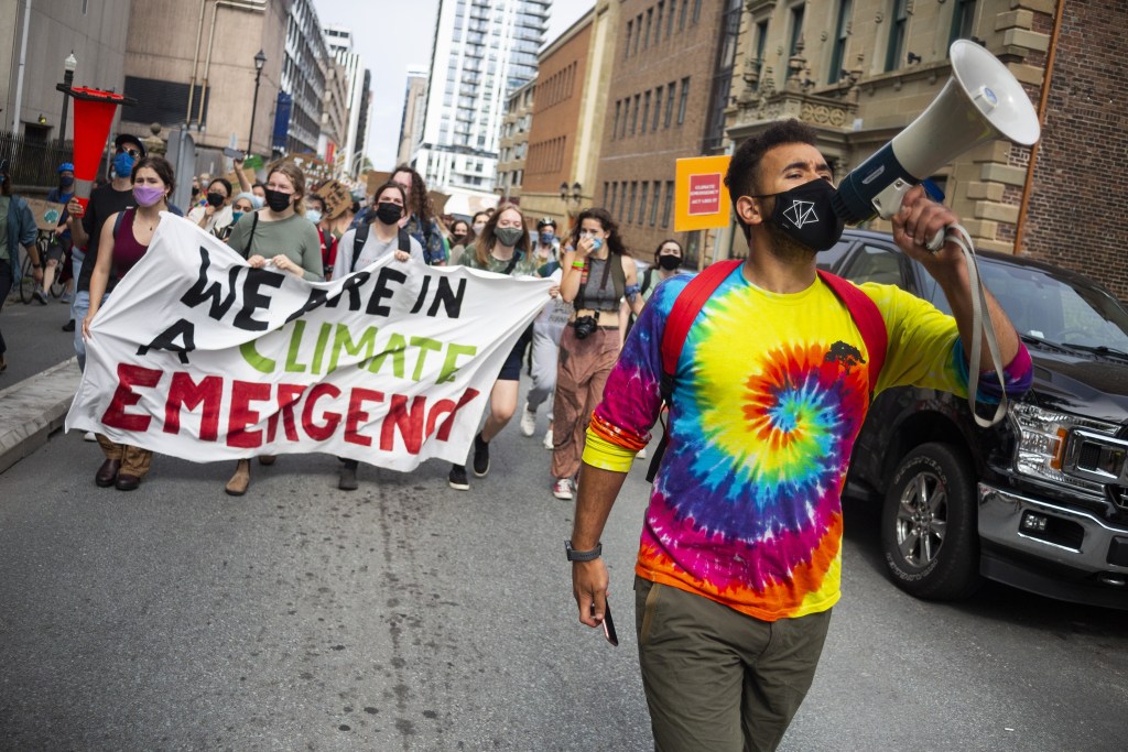A man wearing a tie dye shirt yells into a megaphone. Behind him, there are hundreds of people marching through a city street hold signs. In the front of the crowd, a few of the protesters hold a banner reading, "We are in a climate emergency."
