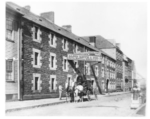 A black and white photo of the former Alexander Keiths Brewery. The building is dark and white brick trim on the windows. A horse and carriage wait outside under the sign.
