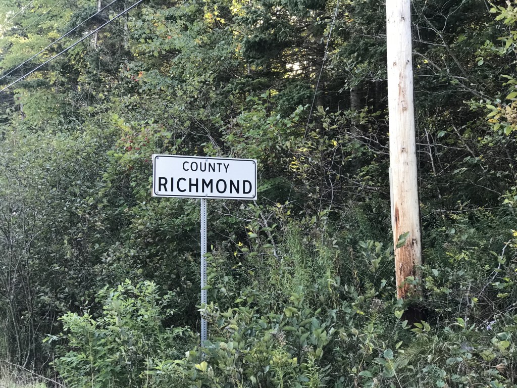 A photo of a white sign that says County Richmond in black font. The sign is next to a wooden telephone pole on the side of the road and surrounded by trees and shrubs.