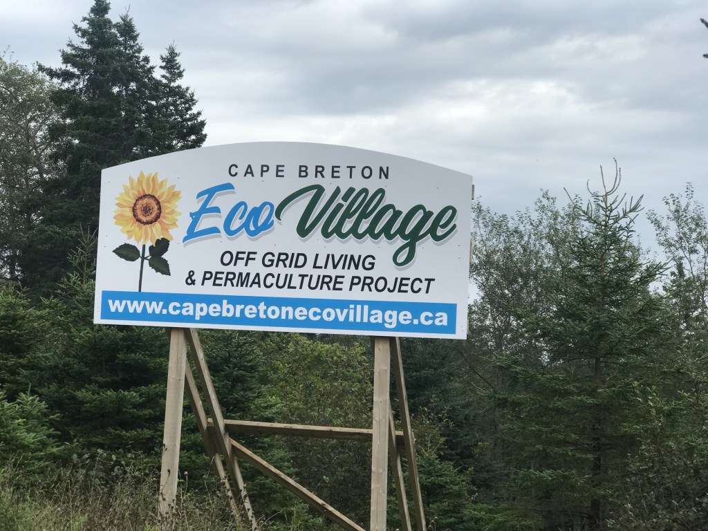 A photo of a sign that says Cape Breton Eco Village Off Grid Living and Permaculture Project. The sign also features yellow flower and a website www.capebretonecovillage.ca