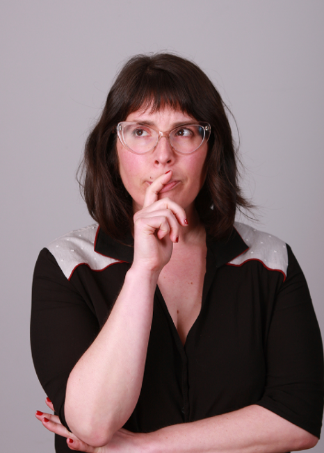 Headshot of a woman with shoulder-length hair and glasses, holding her hand with index finger pointing upward in front of her mouth. She is standing in front of a neutral background.
