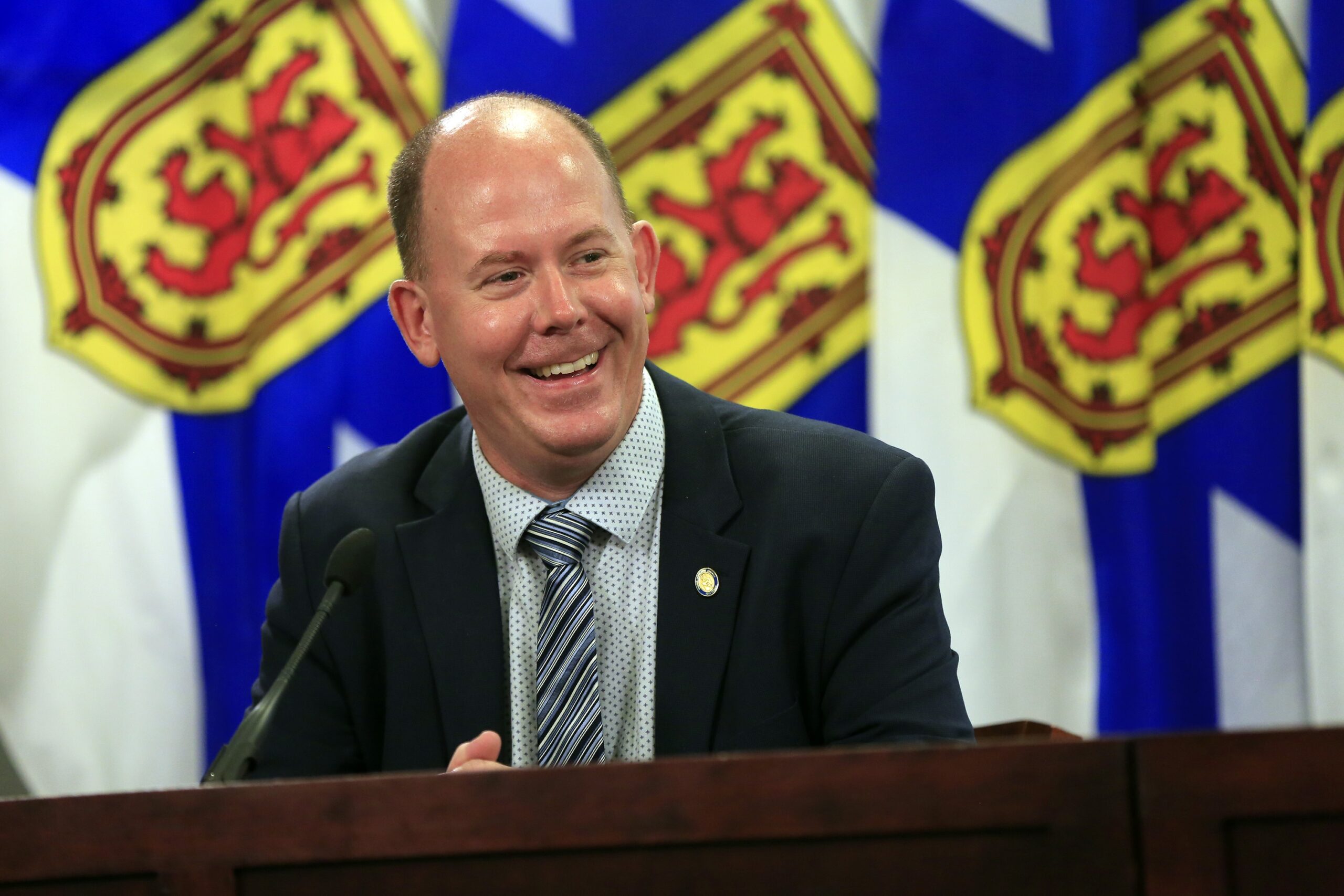 A man wearing a dark suit with a polka dot shirt and striped tie laughs at a podium. In the background are three Nova Scotia flags, coloured blue, yellow and red.