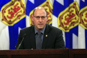 A man wearing a black suit, grey shirt and grey tie speaks into a microphone at a podium. In the background are three Nova Scotia flags, coloured blue, yellow and red.