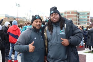Auburn Eagles head football coach Dion Thomas-Hodges smiles for the camera and gives thumbs up(left) with assistant coach Terrence Mendes. The two are wearing grey hoodies, black toques, and are standing on the field in Huskie Stadium with a crowd of fans behind them. 