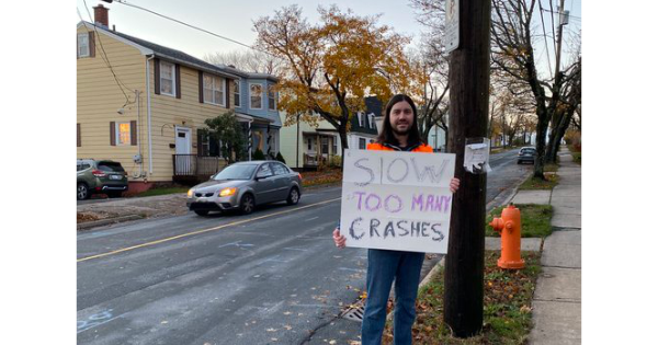A photo of a young man with long hair, an orange jacket, and jeans standing on the side of a street holding a sign that says Slow Down Too Many Crashes.