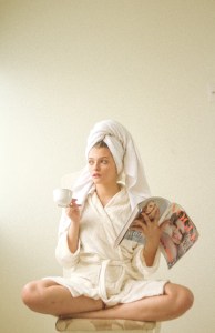 A woman with her hair wrapped in a white towel and wearing a white robe sits crossed legged on the floor with a cup of coffee in one hand and a magazine in the other.