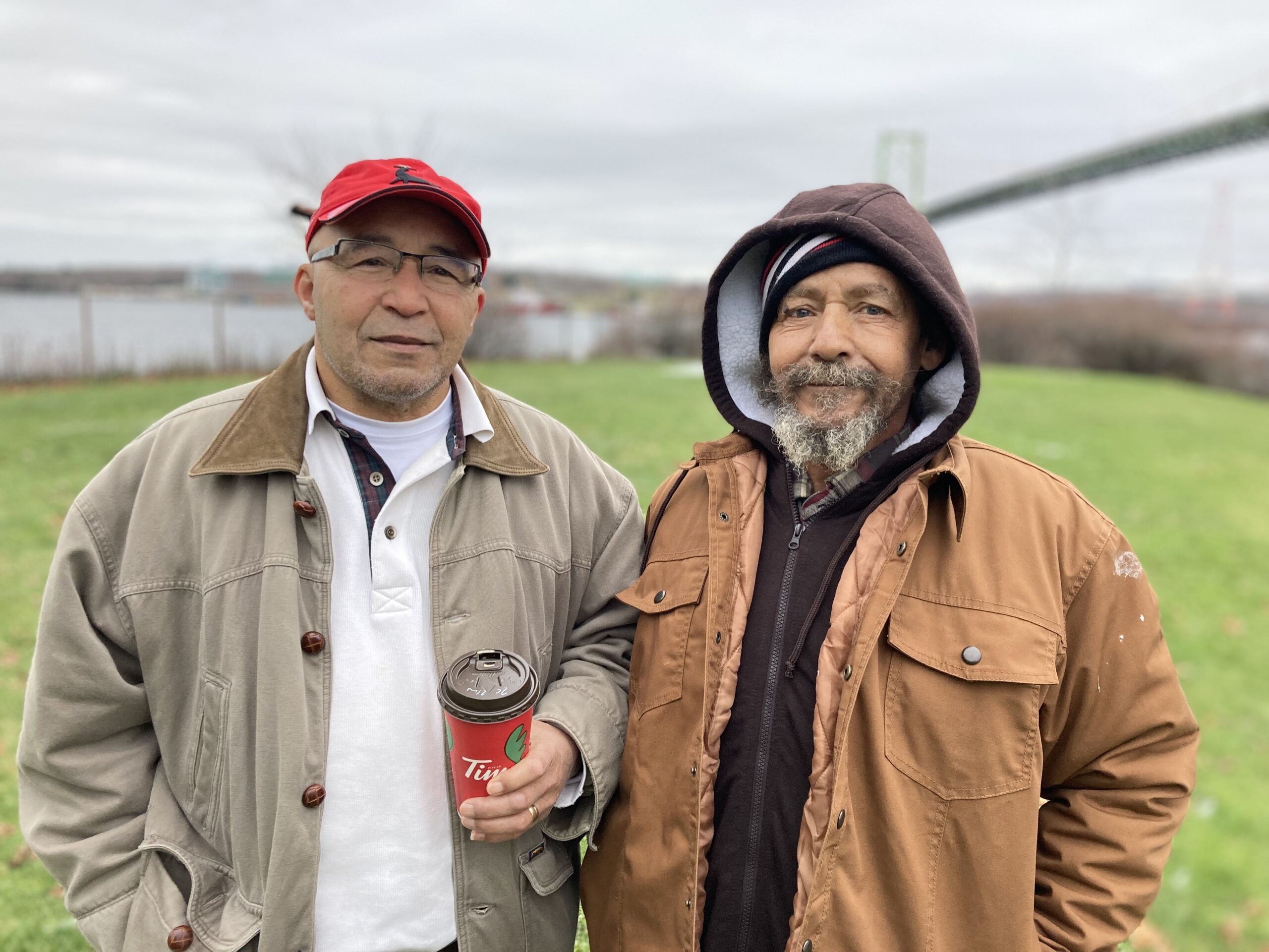 Black man with tanned fall jacket, red hat, and Tim hortons cup, stands next to Black man in brown hhoding and lighter brown coat with the MacKay Bridge in the background blurred out