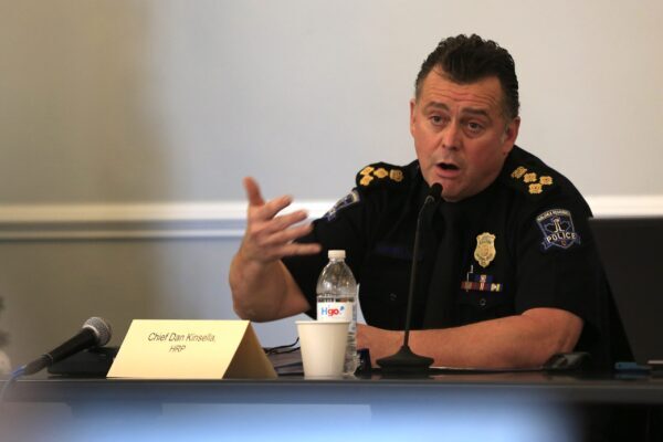 A man in a black uniform speaks while gesturing with his right hand. On the uniform is the Halifax Regional Police logo, along with a badge and insignia denoting rank. On the table in front of the man is a name plate, a microphone and a water bottle. The background is grey.