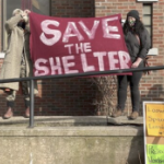 Two people on a set of steps at a brick building hold a red banner which reads "save the shelter"