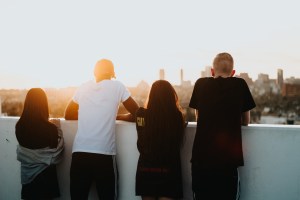 Four young people stand on a rooftop looking over a city where the sun is setting. 