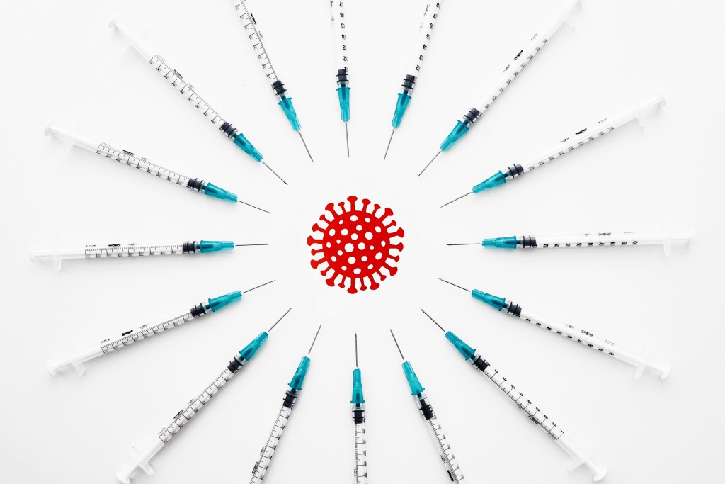 Illustration of a red coronavirus, with distinctive spikes, surrounded by syringes arranged in a circle, with the pointy tips aimed at the stylized virus.