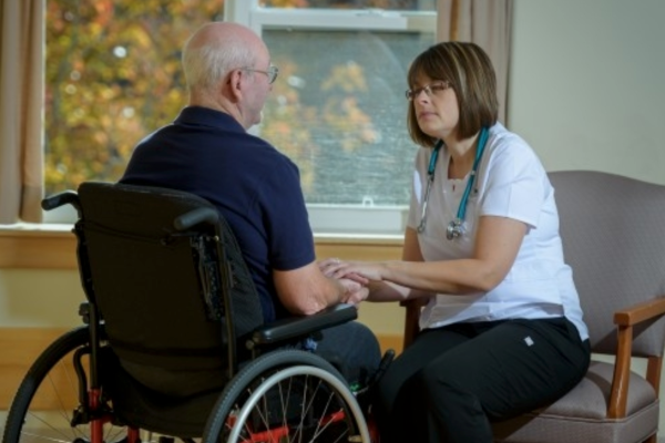 A white woman dressed in nurses' gear holds the hands of a white senior man in a wheelchair as they chat in a long term care facility