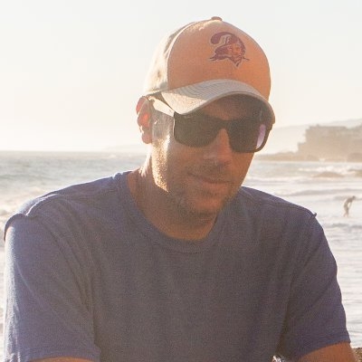 Slightly faded image of a white man in sunglasses and a ballcap, with open water behind him.