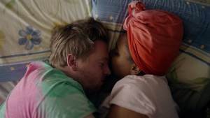 A still from a film that shows a white man and a Black woman lying in bed.