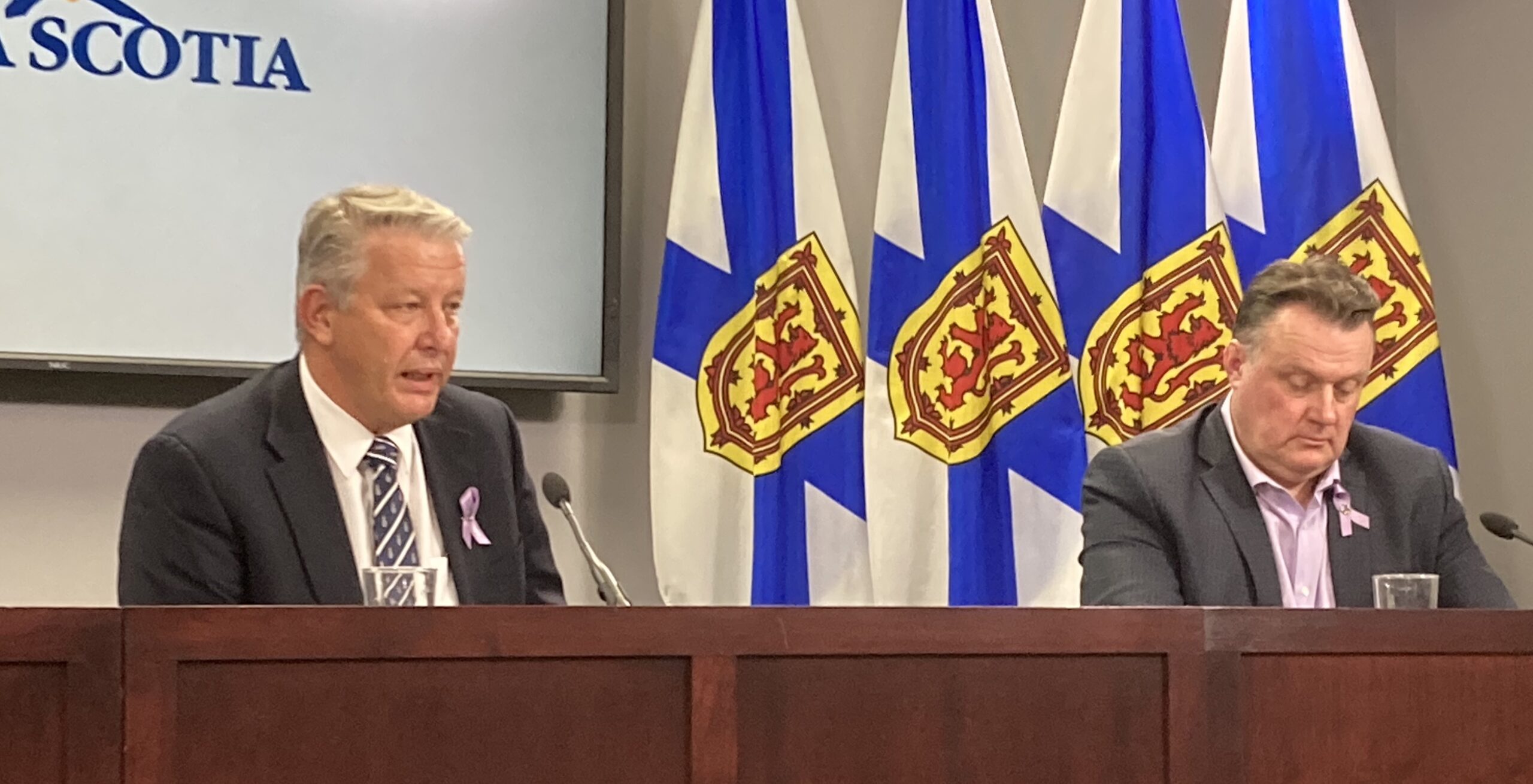 Two men sit at a desk with Nova Scotia flags in the background