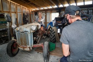 A young blond woman sits on an old rusty tractor in a shed. In the foreground, a camera operator looks through a camera viewfinder, filming the woman.