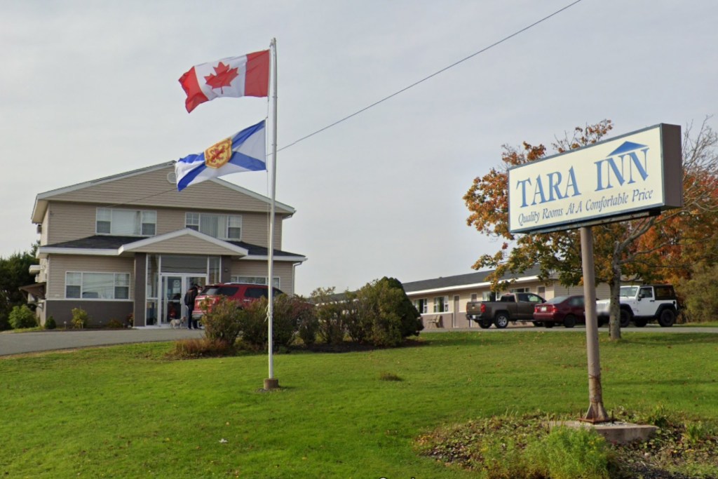 A motel is seen on an overcast day. In the front lawn, there's a flag pole with the Canadian and Nova Scotian flags flying. In front of the flag pole is a sign reading, TARA INN Quality Rooms At A Comfortable Price."
