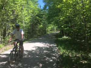 A white man with white stubble sits on his bicycle on a bike trail. The trail is gravel and surrounded by green foliage. It's a sunny day