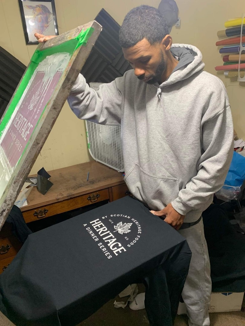 A black man in a gray hoodie presses a t-shirt with the word "Heritage" above