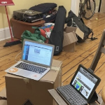 Two laptops sit on a cardboard box and a chair in an apartment with other boxes of household goods