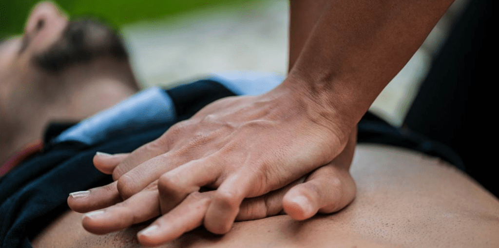 A close up of a person's two hands performing chest compressions on a bearded man on the ground.