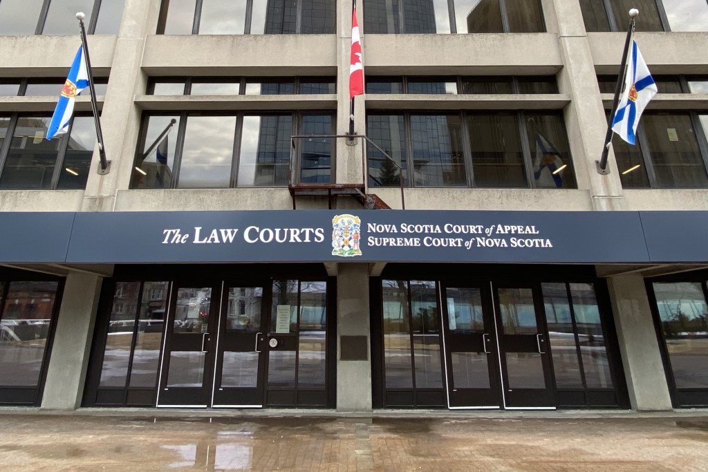 A concrete building is seen on a grey day. The sign says "The Law Courts, Nova Scotia Court of Appeal, Supreme Court of Nova Scotia." There are three flags — two Nova Scotian and one Canadian, in the centre.