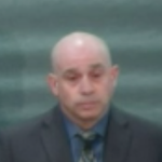A bald white man testifying in front of a dark grey green wall