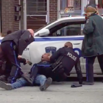 A Black man, Clinton Fraser, is pinned down on the street, held down by a white cop with three other white cops over him, one holding a taser