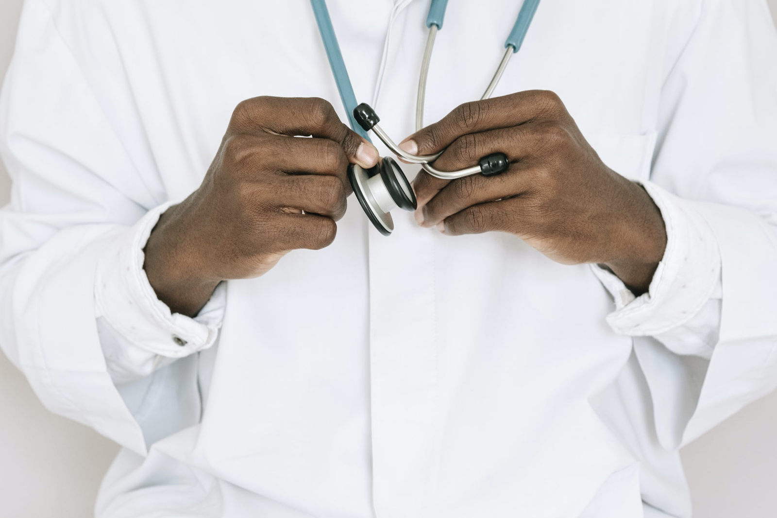 A doctor in a white coat grips a stethoscope in both hands in front of him. The photo is taken from the neck to hips.
