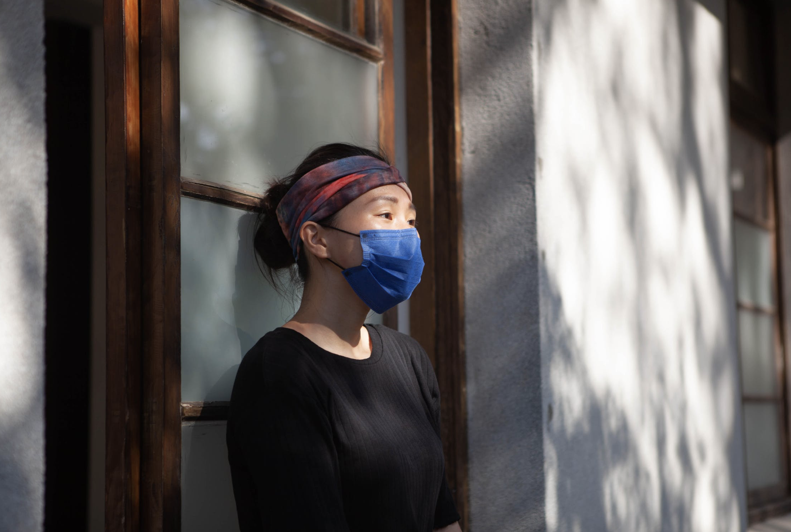 A woman wearing a black long sleeved shirt and a blue mask stands in front of a door way against a concrete building with light filtering on her face.