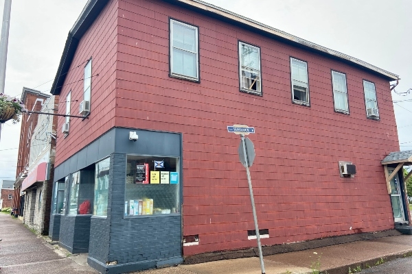 The exterior of a two-storey building with red siding and a green storefront. The building is on a corner lot in a downtown area. 