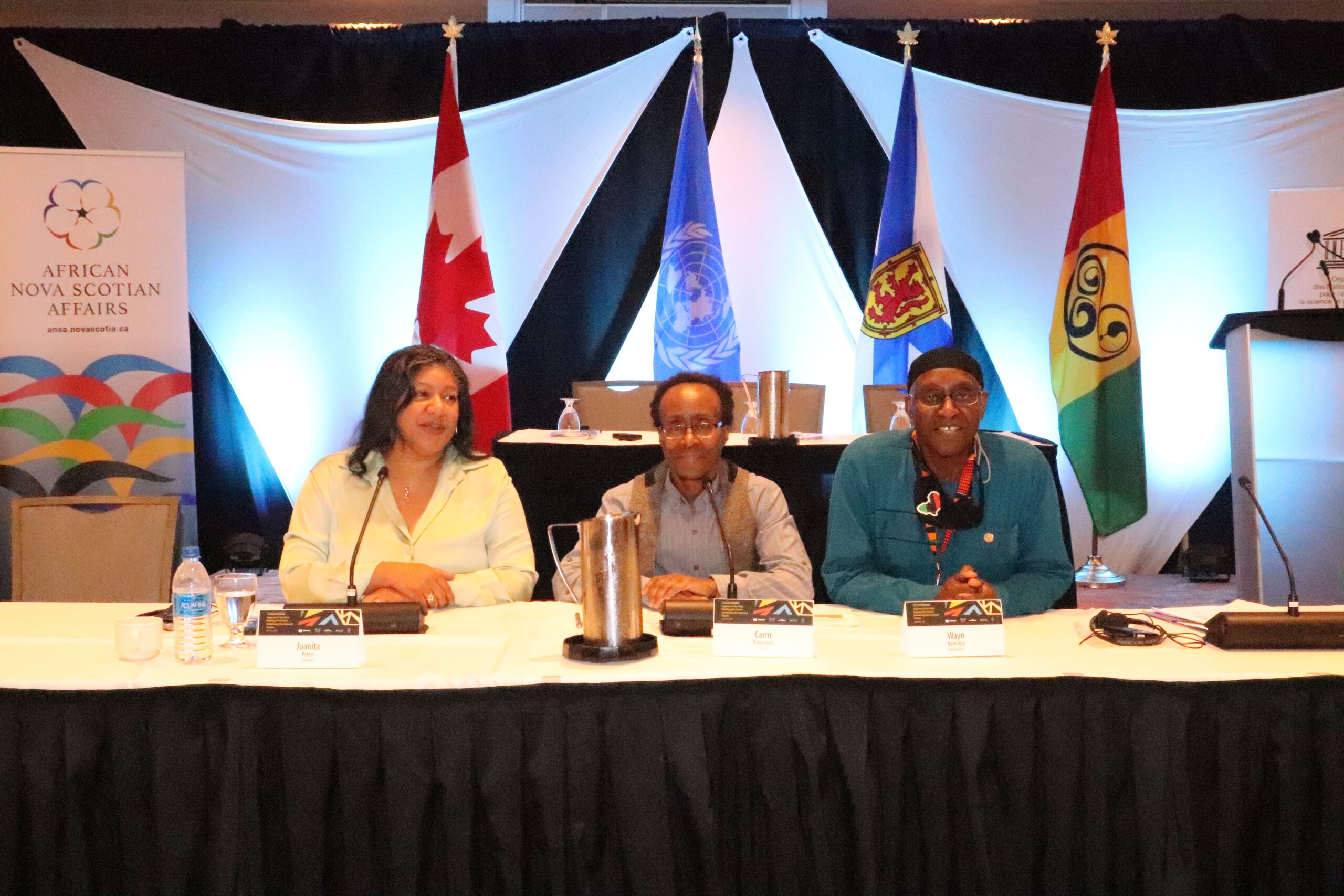 Canadian flag, UN flag, Nova Scotian flag, and African Nova Scotian flag in the background as three Black Nova Scotian sit at table for a panel discussion.
