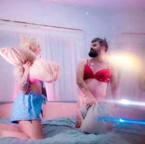 A blonde woman and a white man with a dark beard, both wearing pajama bottoms and either a red or a pink bra, have a pillow fight on a bed.