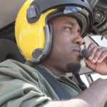 A Black man in a yellow helmet flying a helicopter